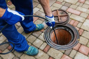 A worker in gloves and blue work clothes inspects a sewer drain with a cable tool on a paved brick surface. The round drain cover lies open beside them, showcasing the professional Plumbing Services by Impact Plumbing in Las Vegas NV.