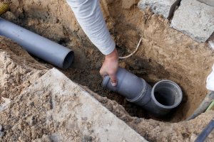 A person’s hand installing gray PVC pipes in a trench, possibly for plumbing or drainage work. Dirt and construction materials are scattered around—typical of professional Plumbing Services in Las Vegas NV, like those from Impact Plumbing.