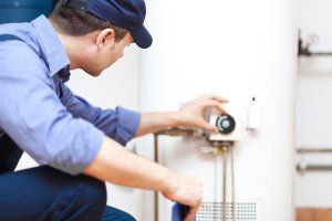 A technician from Impact Plumbing, wearing a blue uniform and cap, adjusts the settings on a water heater in a utility room, delivering expert plumbing services in Las Vegas NV.