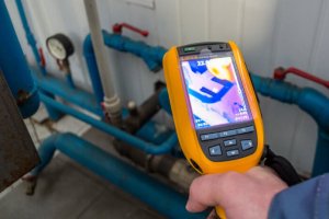 A person holds a yellow thermal imaging camera pointed at blue pipes; the display shows a heat map of the pipes, highlighting temperature differences—showcasing advanced plumbing services by Impact Plumbing in Las Vegas NV.