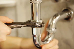 A person uses a wrench to tighten or loosen a shiny, chrome pipe under a sink, suggesting plumbing repair or maintenance work by Impact Plumbing in Las Vegas NV.